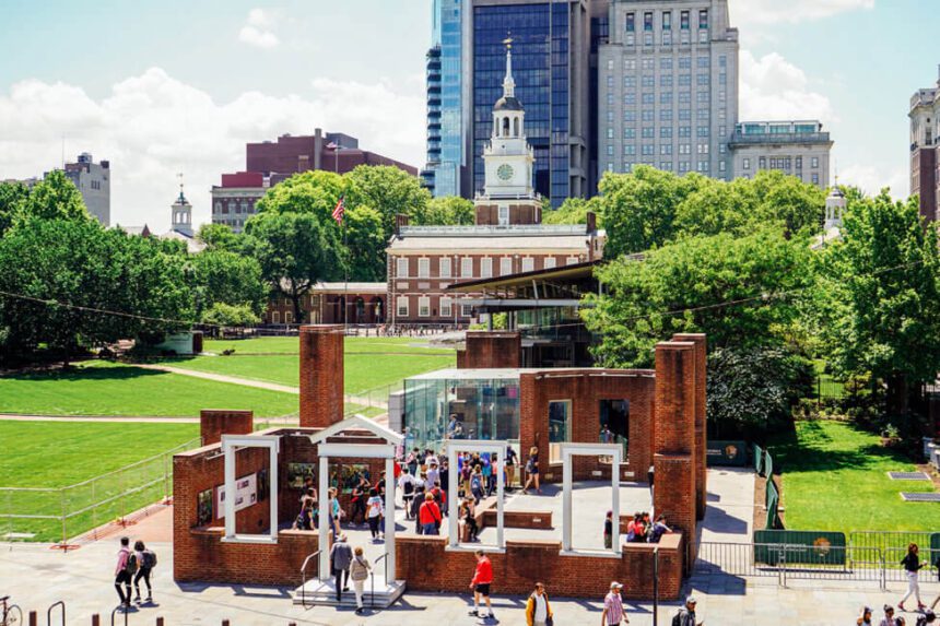 Independence Hall, a striking brick and white building appears in the distance. In front, there is the Presidents' House, a historic site filled with visitors. Lush, green lawns surround the buildings, with green trees off to the right and to the left. The sky above is bright with fluffy clouds.