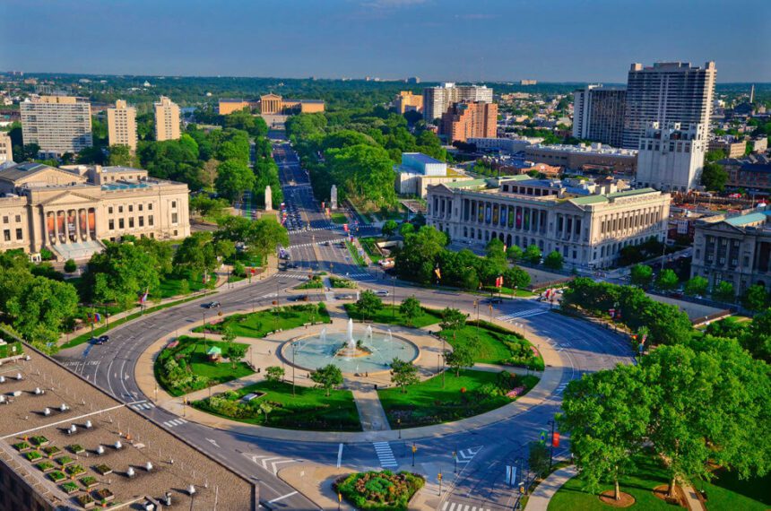 This is a bird's eye view of the Benjamin Franklin Parkway. There is a large fountain in the center of a circle. There are green trees around it. There are bright green trees spread along the highway. There are large buildings on either side. The sky above is a darker blue, almost gray. There are cars shown on the road.