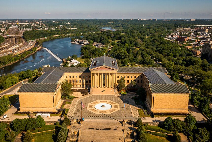 A large museum is shown. There is a fountain in front of it. Two flag poles are shown on either side of the fountain. There is a river shown beyond the structure, as well as green trees surrounding it. The image is taken from above so the angle appears to be looking down, as a sort of bird's eye view/aerial view.