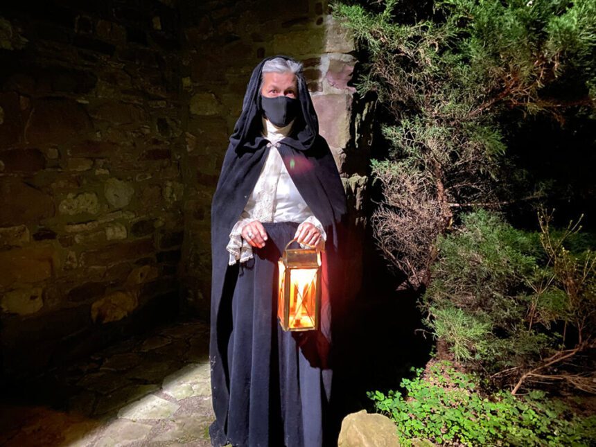 A woman dressed in costume stands holding a lantern. It is dark outside. A tree is behind her. The lantern glows a yellowish orange.