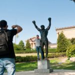 A man is shown wearing a backpack taking a picture of another man who is posing with a large statue of Rocky Balboa, a fictional boxer. The statue shows Rocky holding his arms up, with his fists in the air. Behind the statue, there is the Philadelphia Museum of Art, a large structure surrounded by lush green lawns and full green trees and bushes.