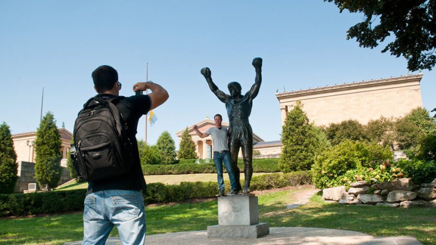 A man is shown wearing a backpack taking a picture of another man who is posing with a large statue of Rocky Balboa, a fictional boxer. The statue shows Rocky holding his arms up, with his fists in the air. Behind the statue, there is the Philadelphia Museum of Art, a large structure surrounded by lush green lawns and full green trees and bushes.