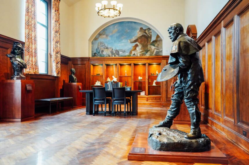 A large sculpture of a man is shown off to the right. There are two busts on pedestals off to the left. in the center of the room there is a table and chairs. The walls are a deep brown wood. The ceilings are a light cream color. There are cases of smaller sculptures lit up.