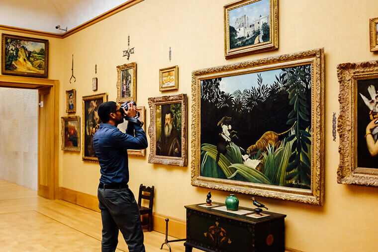 A man is shown taking a picture of a painting with a camera. There are multiple paintings shown hanging on a yellowish gold wall. 