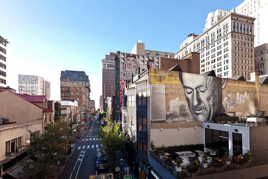 A mural of a man looking down is shown on the side of a building on the right. There appears to be a rooftop patio area right beneath the man in the mural's gaze. To the left, there is a street shown below. There are multiple cars shown on the road. There are buildings off to the left. The sky above is clear of any clouds and is a light blue, almost white on the left.