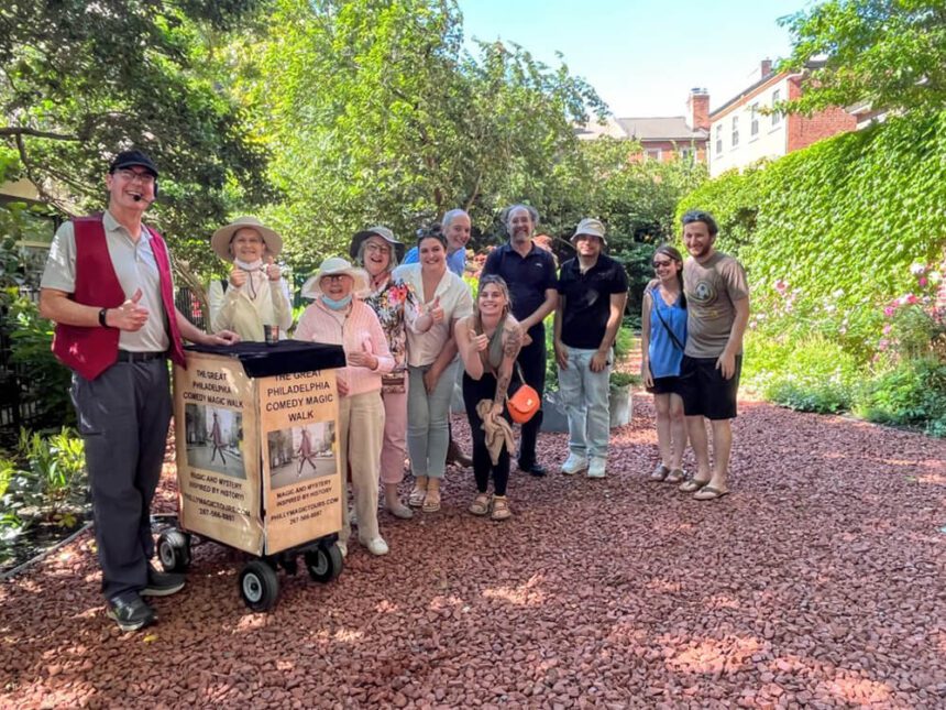A group of people stand with a tour guide. There are lush green trees and bushes behind them. They stand on a gravel path. The tour guide has a cart in front of him that reads The Great Philadelphia Comedy Magic Walk.