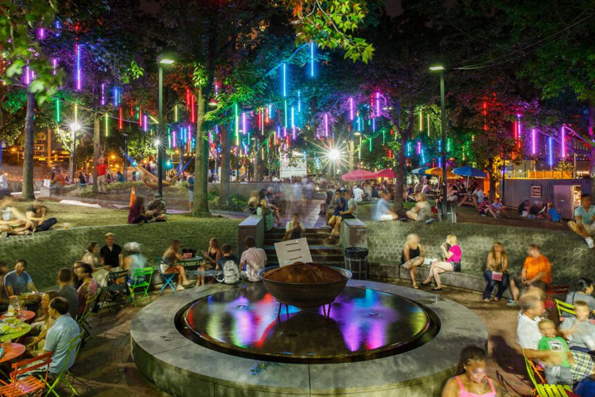 A vibrant outdoor space is shown. There are brightly colored lights hanging from trees throughout the area. There are people shown sitting on colorful chairs and around tables. There are hammocks set up. It is nighttime and the park is full of color and life.