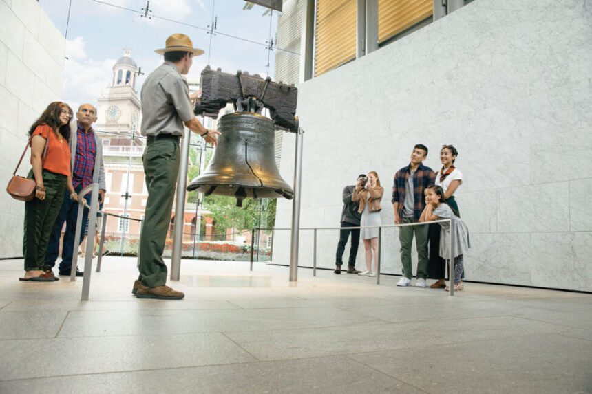 A man stands in front of the Liberty Bell, he appears to me speaking to the people standing on either side of the large bell. There are ropes around the bell to prevent from visitors from getting too close. There is a large glass window behind it letting in natural light. Independence Hall is shown on the other side of the window.