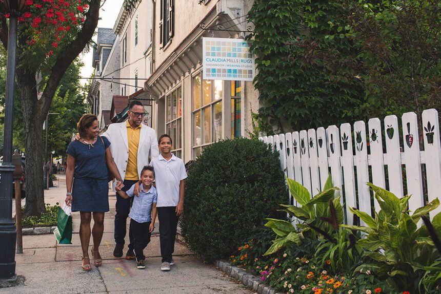 A woman, man, and two young boys are shown walking down a quiet street. There is a bush to the right, along with colorful flowers lining the walkway. There is a white picket fence with artwork at the top. Beyond the family walking, there are storefronts in Chestnut Hill.