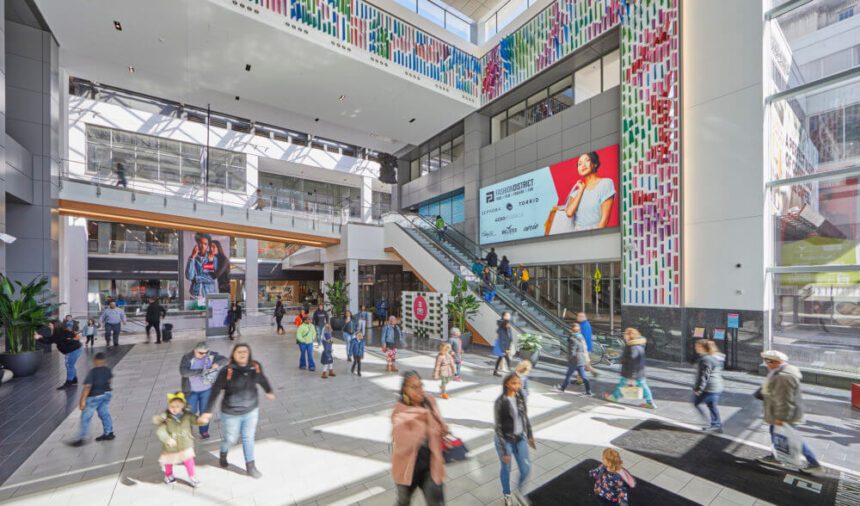 A shopping mall is filled with people walking around. There are people going up the escalator to the right. Large posters and billboards are thrown throughout the indoor space.