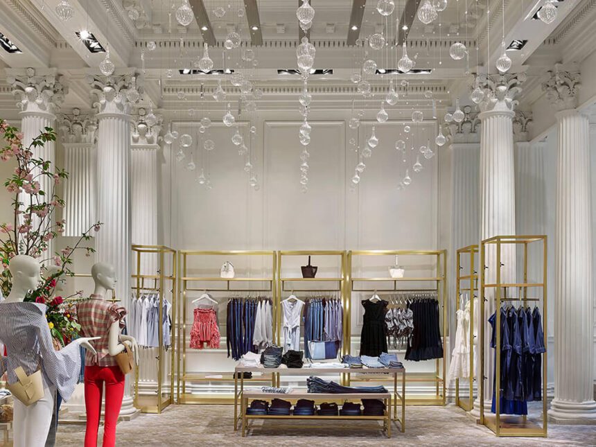 An open space is filled with gold clothing racks with fashionable wardrobe pieces. Two mannequins are off to the left, one is wearing bright red pants, the other is wearing white pants. Clear light fixtures and chandeliers hang from the ceiling overhead.