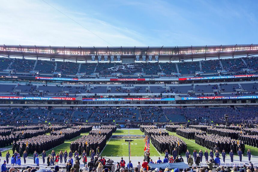 There are rows of officers representing the Army and Navy on the field inside of Lincoln Financial Field. The sky above is a light blue on the right, and white on the left. There are fans in the stands. There are people lining the sidelines of the field.