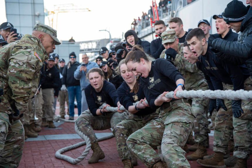 A group of women are shown participating in a tug-of-war competition. There are people on either side cheering them on. Some people shown in the background appear to be recording the event on cell phones.