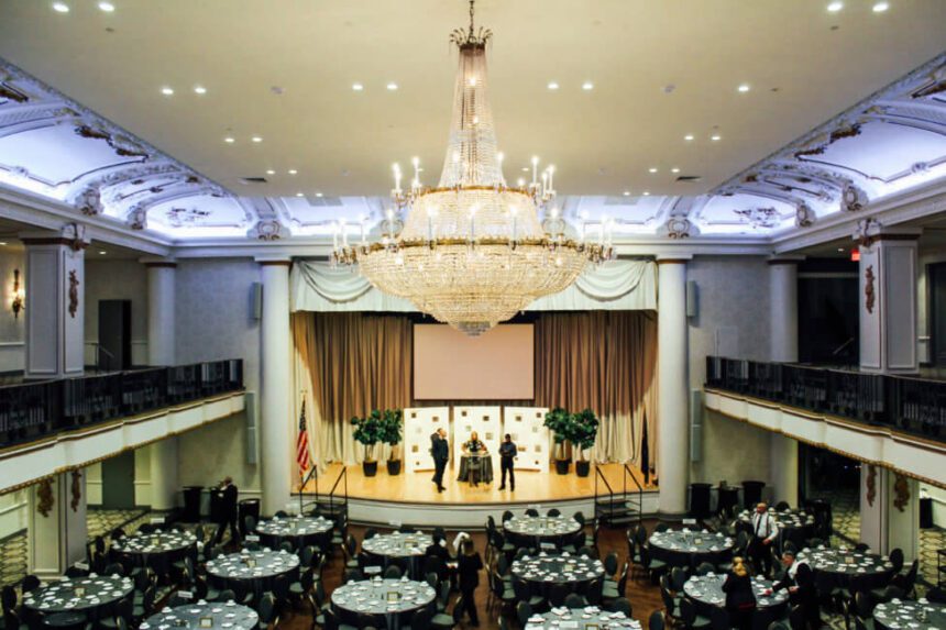 A large room is shown. There are round tables spaced throughout the room. The tables are covered in black table linens with white napkins placed at each setting. There are black chairs around the tables. There is a stage set up with a podium. It is well lit. There is a huge chandelier hanging from the ceiling.