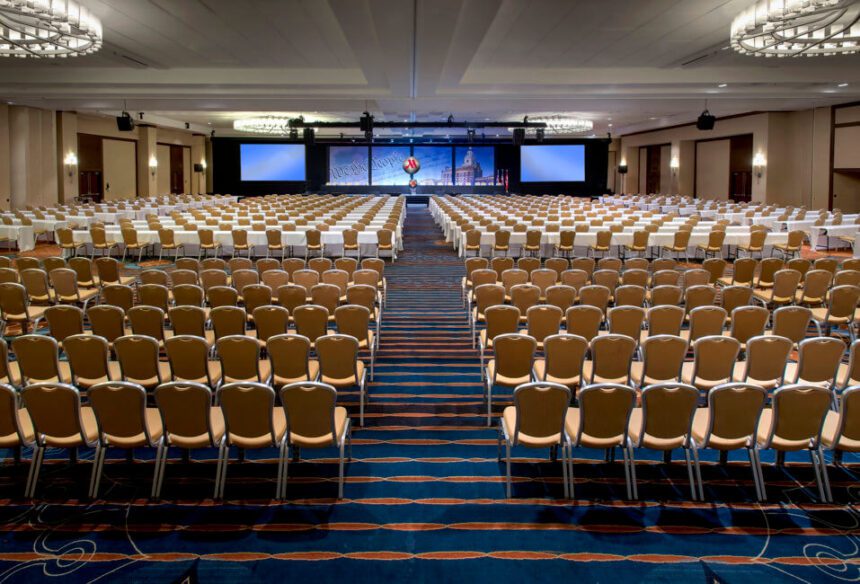 Multiple rows of yellow and silver chairs are lined up. There are more yellow and silver chairs pushed up against white tables in front/ There are large TV screens on either side of the center stage up front. 