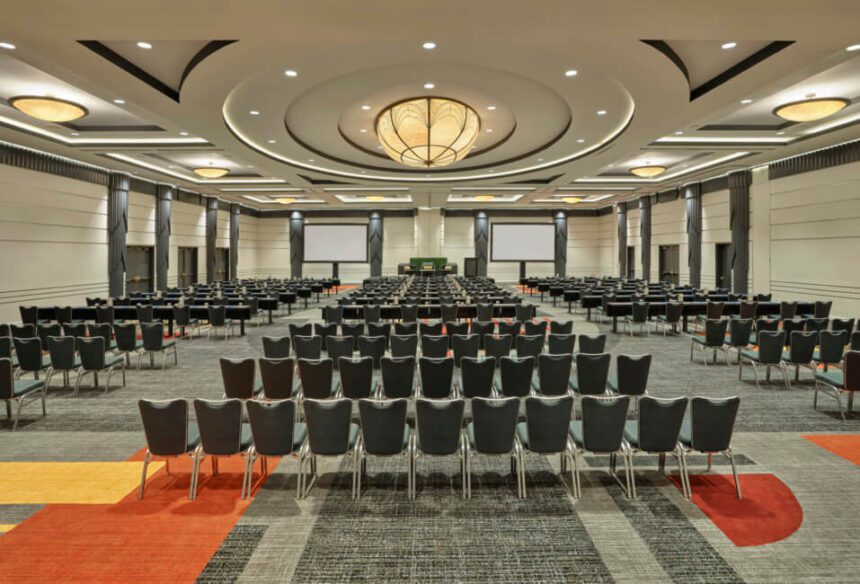 There are several rows of gray and silver chairs shown facing a table in the front of the room. There are large TV screens on either side of the main table up front. The carpet is gray, orange, and yellow.