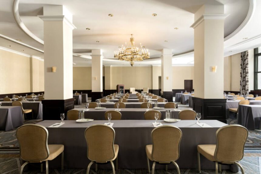 A large room is shown with gray tables set up throughout the space. There are gold and silver chairs set up at each table. There is a gold chandelier hanging from the ceiling. There are four large white pillars shown around the chandelier. Windows on the right are letting in natural light.