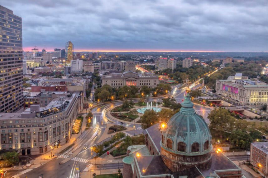 A large mint green dome of a church is shown with a gold crucifix on top of it. Beyond that, there is a fountain in the center of a circle with roads around it. There are large buildings shown throughout. The sky above is a light blueish gray. Street lights are glowing throughout the space