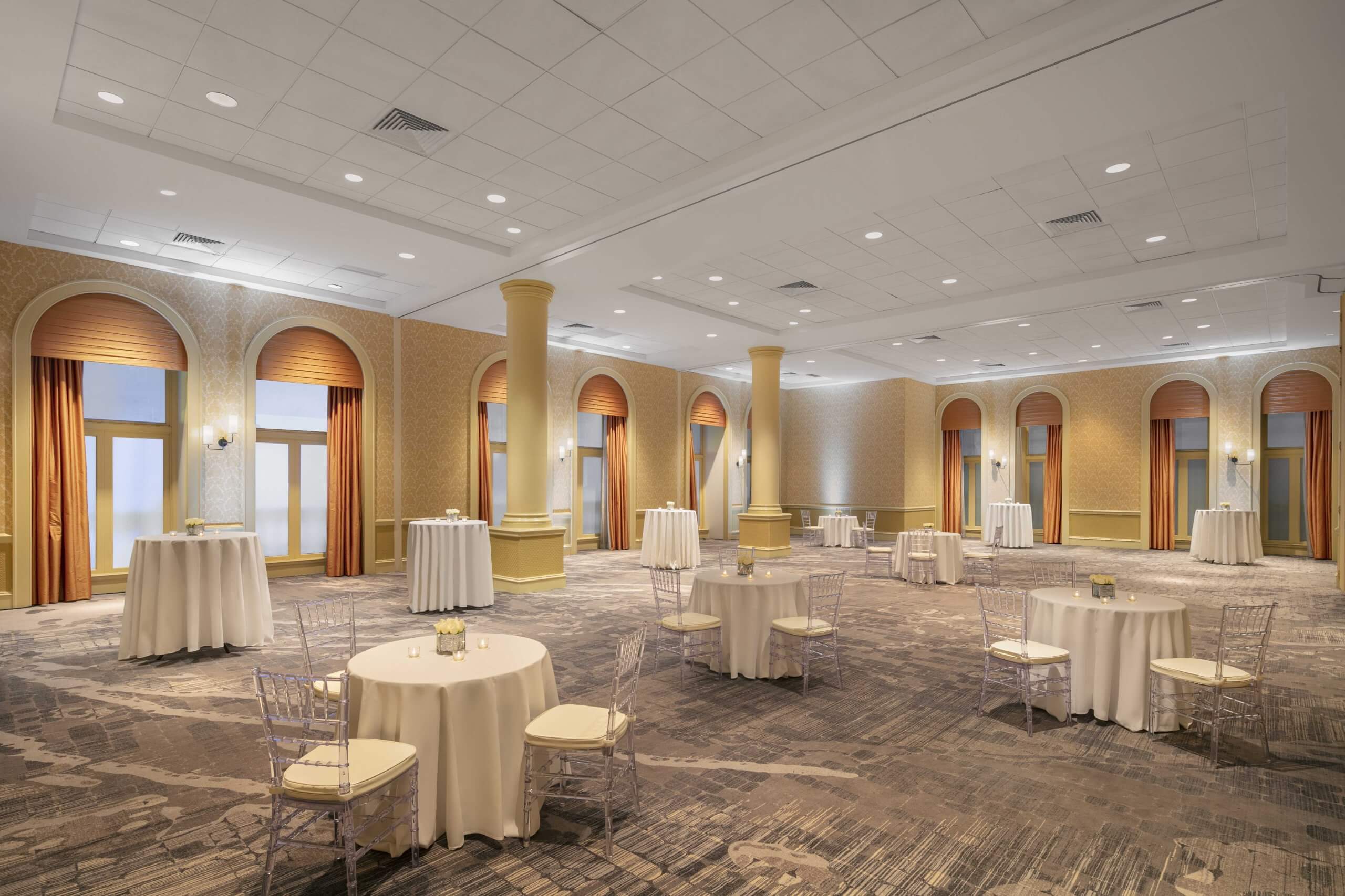 A large ballroom with tables and chairs spread throughout the space. Walls are yellow. Orange curtains hang beside large windows, letting in natural light.