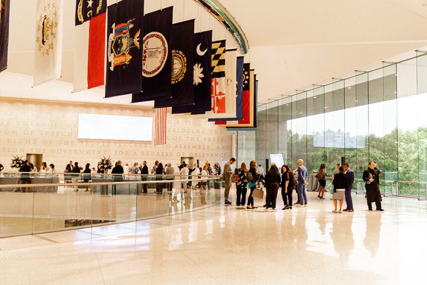 There are large flags hanging down from the ceiling. A large group of people are shown mingling throughout the space. There are tables set up with floral centerpieces. There are large glass windows to the right letting in natural light. Outside, there are green trees.