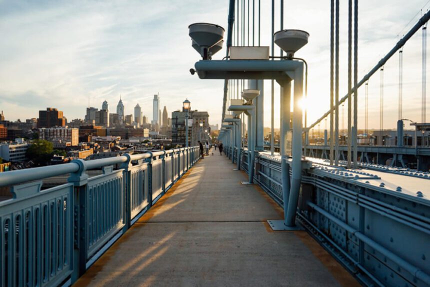 A bridge is shown with a pedestrian walkway. The Philadelphia skyline is shown off to the left. The sun appears to be rising to right beyond the bridge.