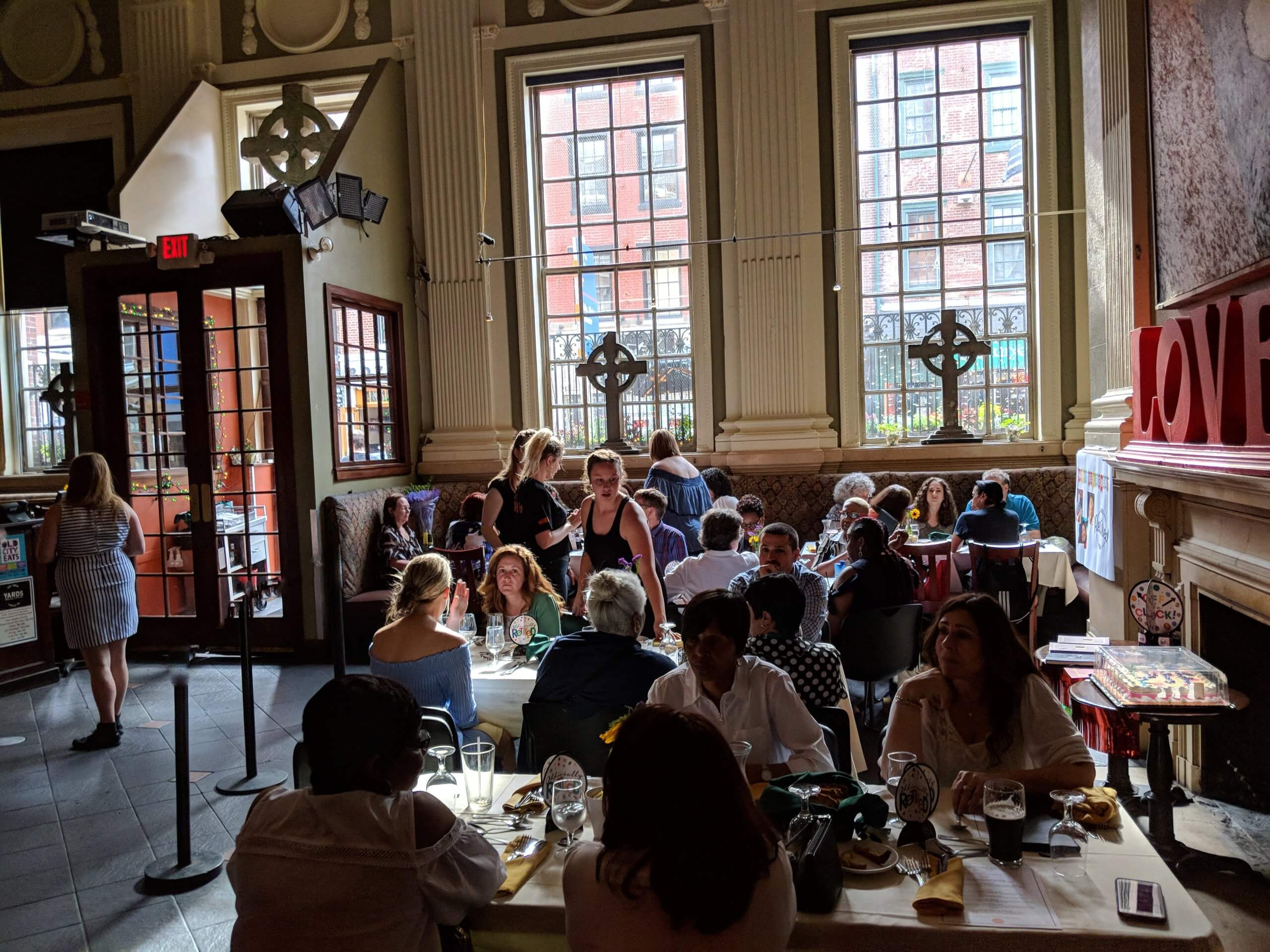 People sit on either side of rectangular tables filling the space. Servers appear near tables, waiting on customers. Red letters spelling "love" sit on a mantle above a fireplace. Two large windows let in natural light. Crosses are propped up on the windowsills.