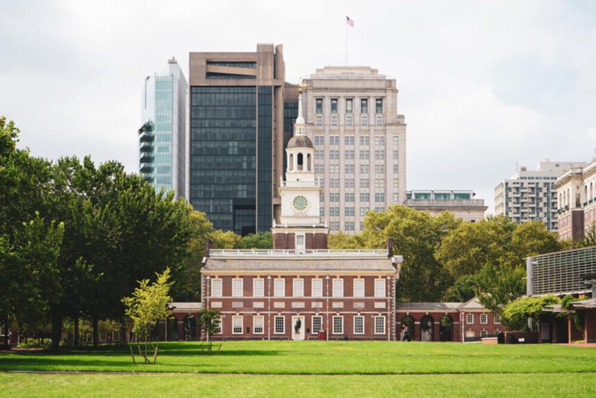 Independence Hall is shown in the center, a lush green lawn is shown in front of it. Trees are shown to the left. The sky above is a bit gray and cloudy.
