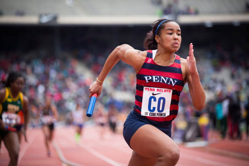 Runner for University of Pennsylvania in blue and red uniform holding a baton races on a track at Franklin Field for the Penn Relays.