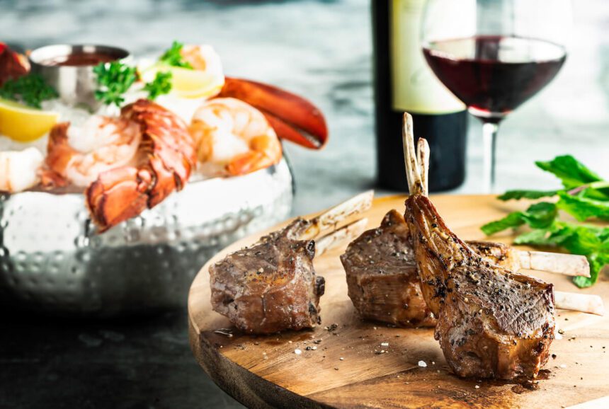 A white dish of shellfish, lemon wedges, and a silver cup of sauce appears off to the left, with pieces of meat on a wooden plate to the right, a wine glass appears behind the plate, along with a bottle of wine