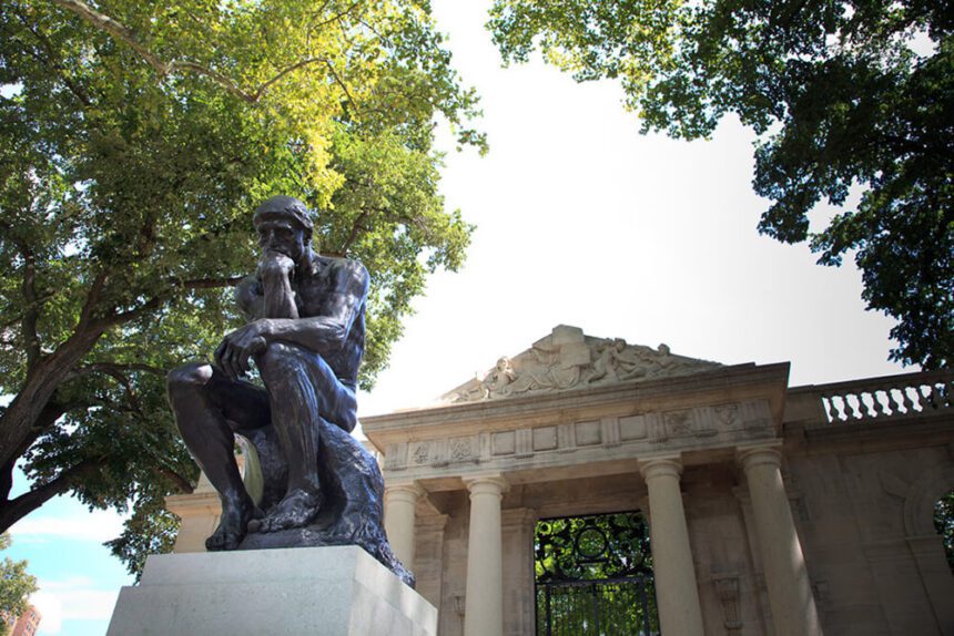A statue is shown outside of a museum. Green leaves from a nearby tree hang overhead.