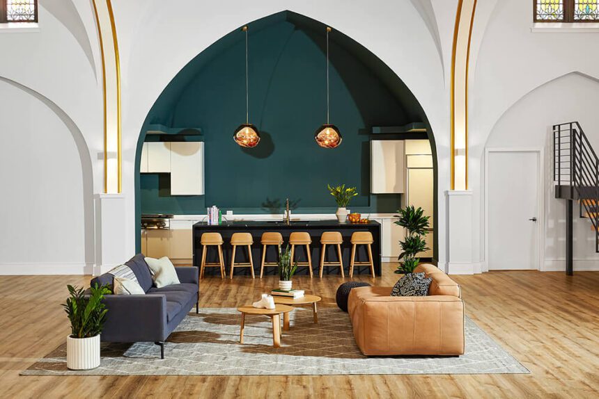 An indoor living space is shown with two couches, one blue, one yellow on either side of a wooden coffee table in the middle. There is a large planter to the left next to the blue couch. Beyond the sitting area, there is a counter with six bar stools pushed up against it. There is a kitchen area beyond it. Two light fixtures hang from the ceiling. A staircase is off to the right.