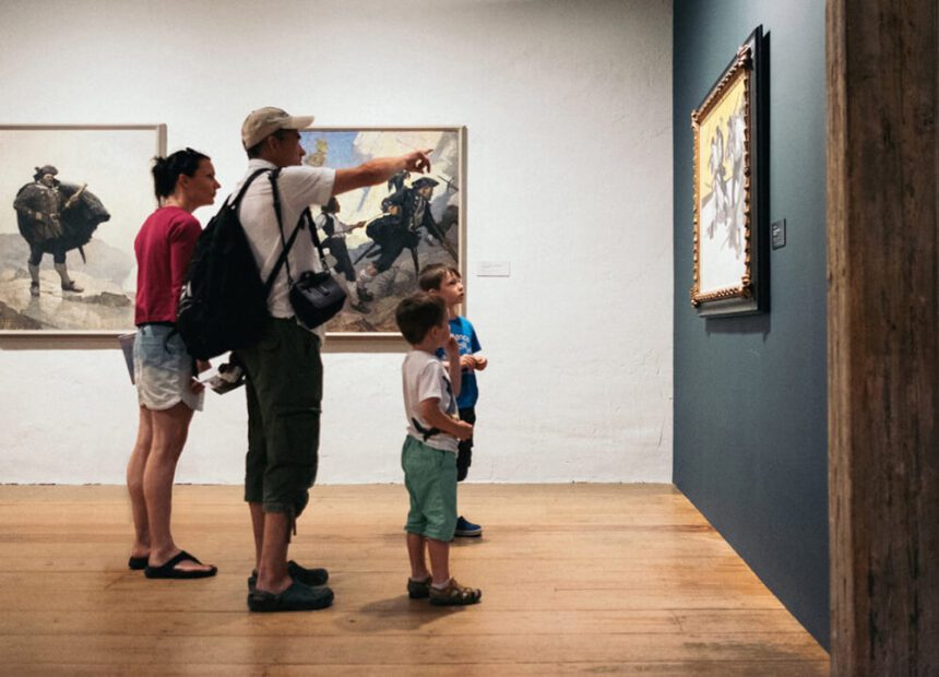 Four individuals stand looking at art. The woman appears to be looking at the painting on the wall, as the man holds his arm out, pointing to the piece. Two young boys stand in front of them, also looking at the piece on the wall.