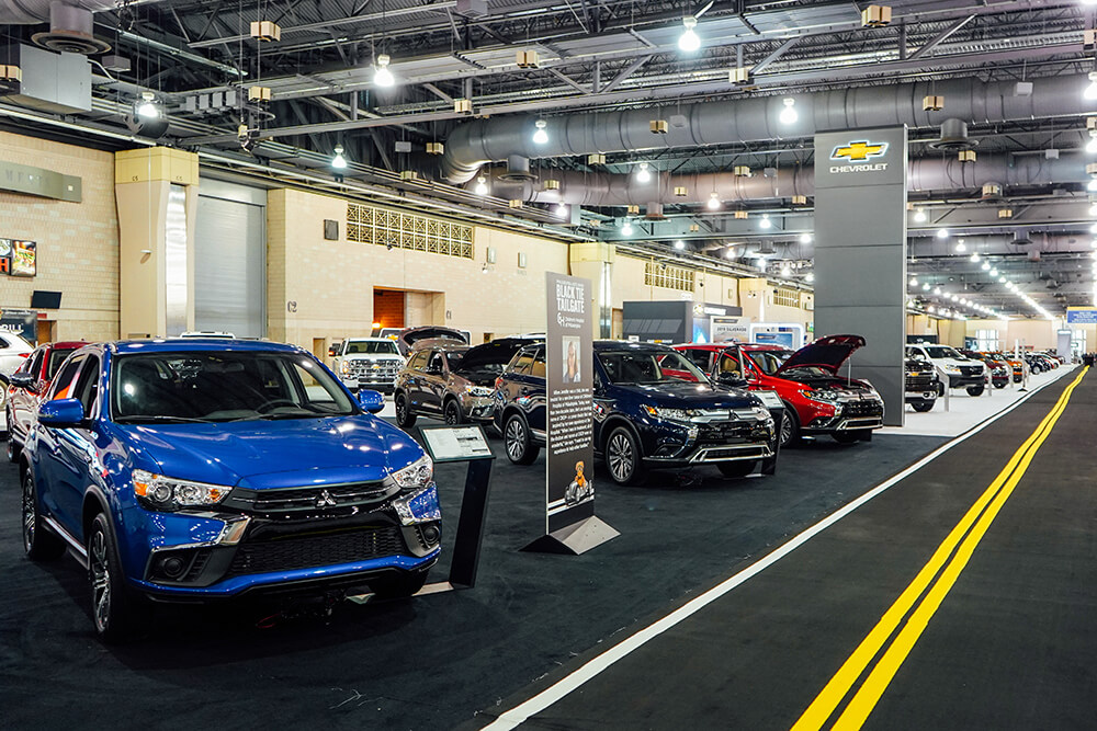 A shiny blue car is off to the left. The carpet inside of the Pennsylvania Convention Center is made to look like a road with the double yellow lines running down the middle. Other shiny cars are on display in a line.