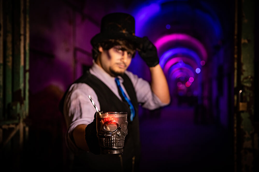 A man holds a drink out in front of him. The glass is shaped like a skull with a red light inside of it. a white and black straw is shown in the glass. The man tilts his hat forward with his other hand. a pink and purple creepy, illusion-inspired hallway appears behind him.
