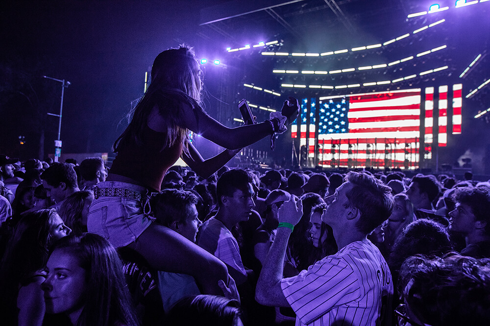 A crowd forms in front of a stage at the Made in America concert, as one woman in the crowd sits on top of a man's shoulders. Another man nearby is wearing a Phillies jersey. An American flag is projected onto the backdrop for the stage.