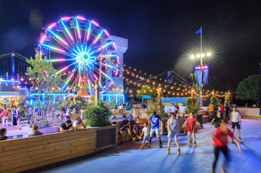 It is nighttime. A large Ferris Wheel is lit up in bright lights off to the left. People are shown roller skating to the right. some people are shown leaning over the perimeter of the rink, watching as people skate by them.