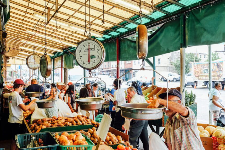 An open-air marketplace is shown. It is bustling with customers picking out their fresh produce. There is a man toward the right using a scale in front of him. There are bins of fresh produce throughout.