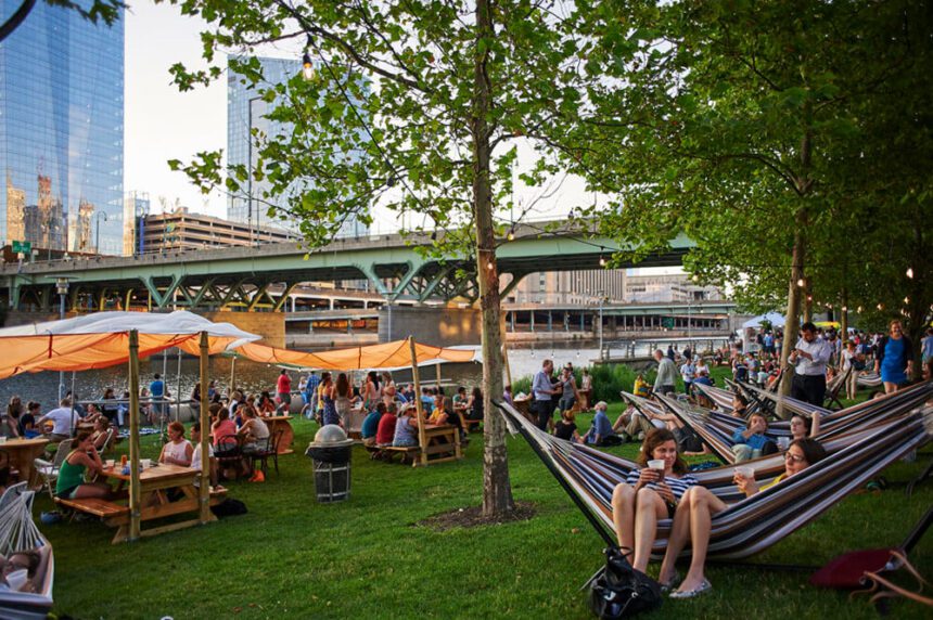 An outdoor park is shown with benches and picnic tables set is to the left with orange drapes overhead covering them from the sun. To the right, there are hammocks lined up. People are shown sitting at the tables and lounging in the hammocks. Some are holding drinks in their hands. Skyscrapers are shown off in the distance to the left.