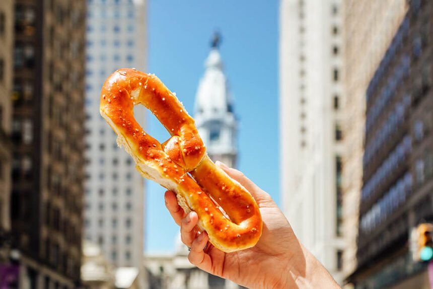 A hand holds a soft pretzel in front of Philadelphia City Hall.