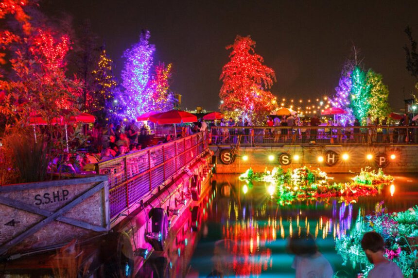 It's nighttime. There are colorful lights illuminating trees. People are shown standing and sitting at tables enjoying food and drinks by the water. Large letters SSHP are shown across the water for Spruce Street Harbor Park.
