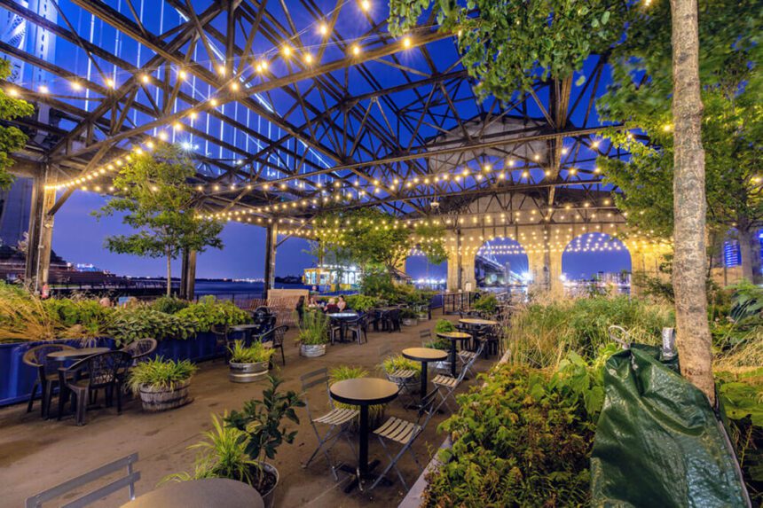 It is nighttime. The sky above is a dark blue. There are lights strung overhead, hanging from the roof of the pier. There are green plants spread throughout the space. Tables and chairs are stationed throughout. A few people are shown off in the back left, sitting around a table.