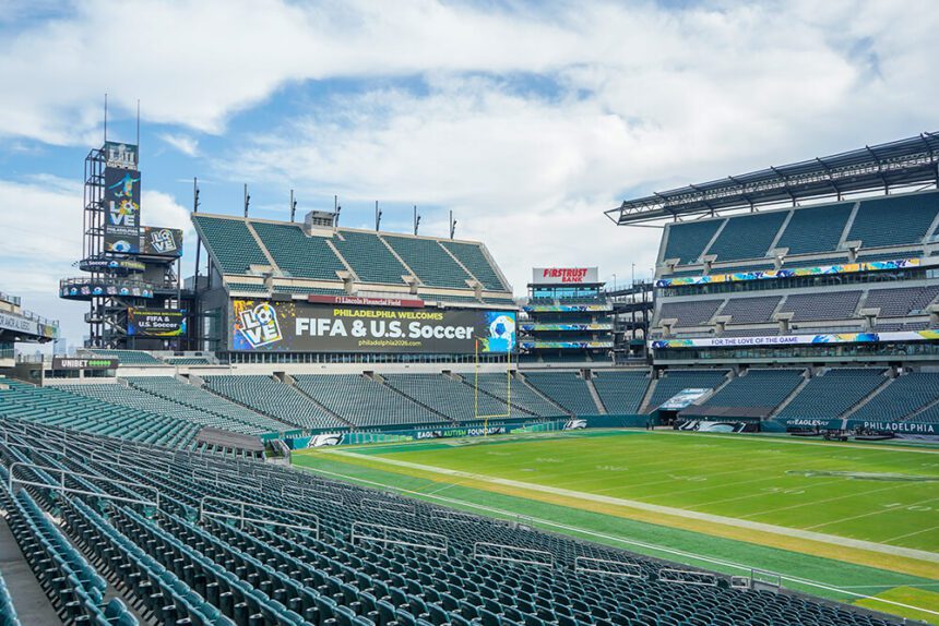 Inside of Lincoln Financial Field, the lush, green field is empty. The seats are empty. The sky above is blue with several clouds. Signage throughout the open-air stadium reads Philadelphia Welcomes FIFA & US Soccer.