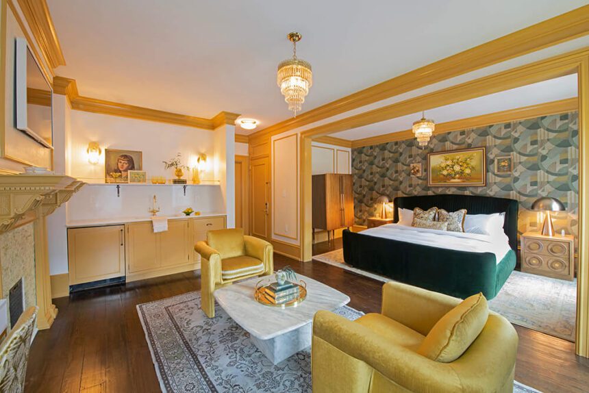 A bright yellow hotel room is shown. There are two yellow cushioned chairs set up on either side of a white coffee table. To the right, there is a green and white bed set up in front of a wall with green and yellow wallpaper. The ceilings are white. Ornate light fixtures hang from the ceiling.