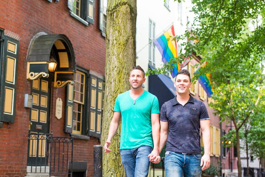 Two men are shwon walking down a street while holding hands. A rainbow flag is shown behind them. A tree is off to the right. A red and yellow building is to the left.