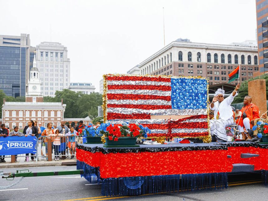 A bright, red, white, and blue float with flowers and other patriotic decor is shown. A woman dressed in all white, waving an African flag in her left hand sits next to an America flag made out of tinsel. Independence Hall is in the background.