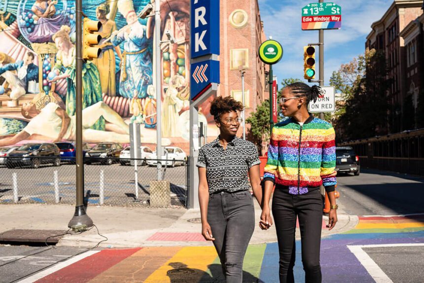 Two women are shown walking across a rainbow crosswalk. The one individual on the right is wearing a zipped up rainbow jacket. A mural is shown behind the individual on the left. A street sign behind them, reads 13th St with a rainbow adorned along the bottom of the sign.