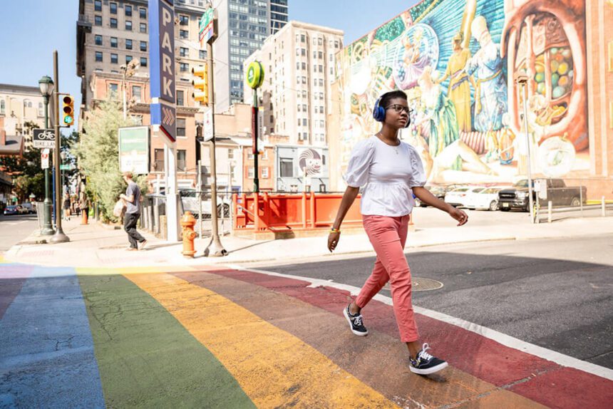 A woman wearing a white shirt, coral pants, and blue headphones walks down a rainbow crosswalk. A colorful is shown off to the right in the distance.