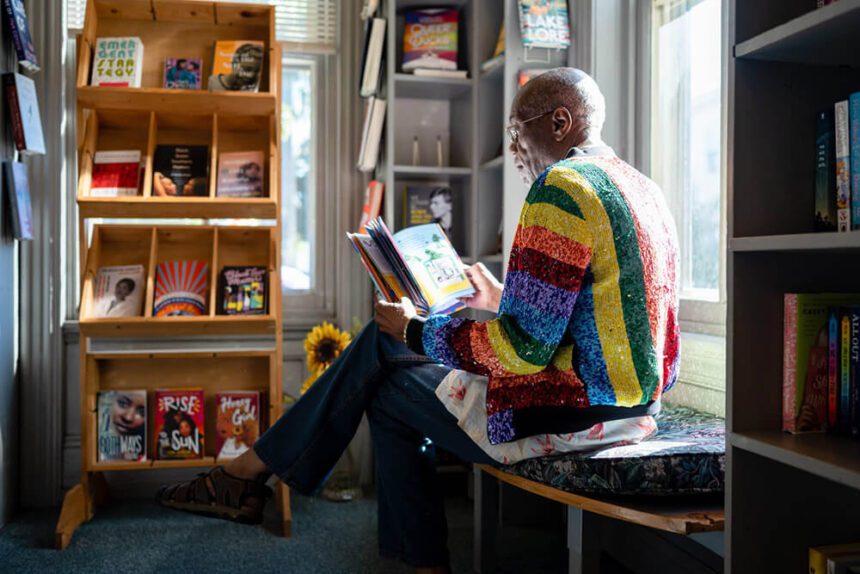 A man wearing a rainbow jacket sits on a bench off to the right. A bookshelf is off to the left. Several books are shown on the shelves. A window behind the man is letting in natural light.