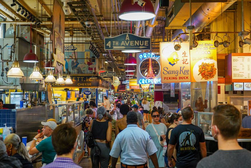 Inside of Reading Terminal Market, people are shown walking past stalls where vendors are selling goods. Bright lights hang from the ceiling above. Menus hang over the stalls.