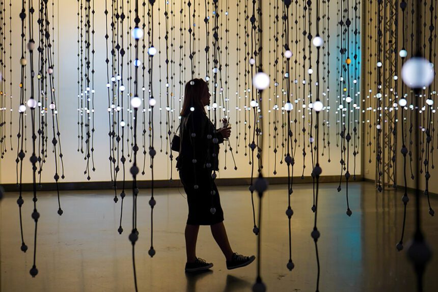 A woman dressed in black is shown walking in a room where there are strands of bright lights hanging as part of an art installation. The lights look like round orbs.